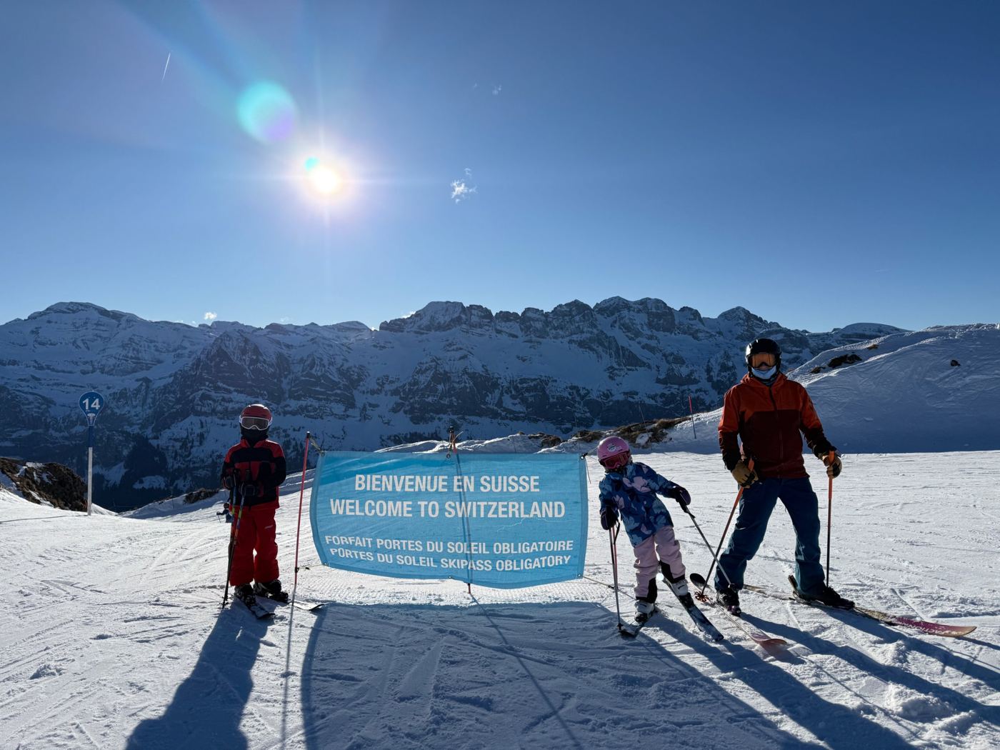 Adam and his two children on skis beside a Welcome to Switzerland banner in the Portes du Soleil